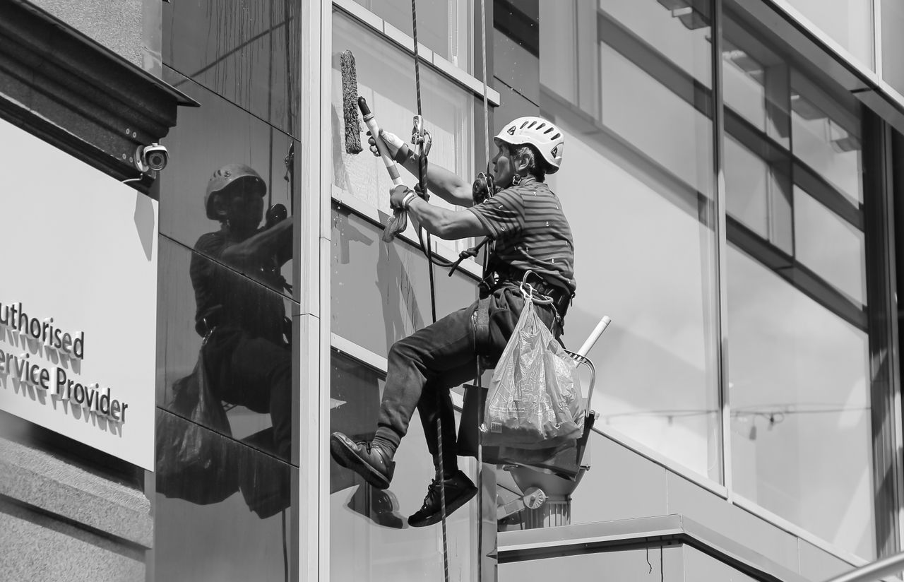services-02 Black and white from below of man steeplejack in protective helmet with professional equipment cleaning glass wall of modern tower