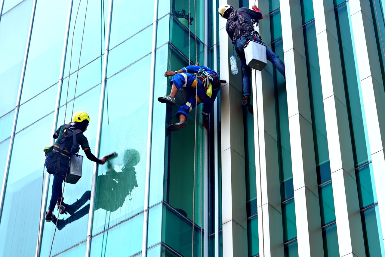 services-01 Three window cleaners suspended on ropes clean a modern glass building facade during the day.