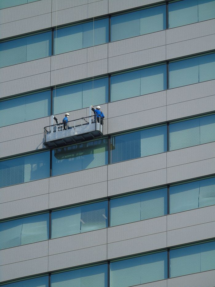 who-we-are Two workers clean windows on a modern glass building facade using a suspended platform.